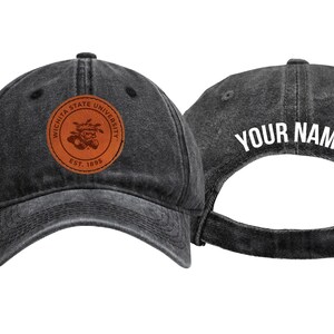 May include: Two charcoal gray baseball caps. One cap features a brown leather patch with the text "Wichita State University" and a logo. The other cap has the text "YOUR NAME" in white on the back. Adjustable strap.