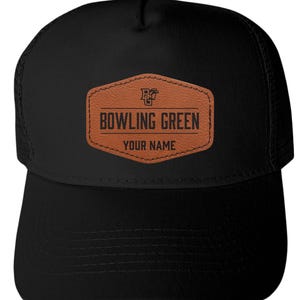 May include: A black trucker hat featuring a brown leather-like patch on the front. The patch displays a logo with the letters BG, the text BOWLING GREEN, and the text YOUR NAME in black lettering. The hat has a solid front panel and mesh back.