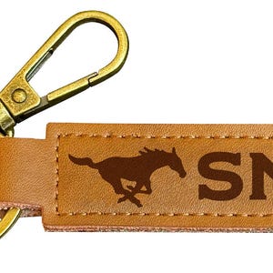 May include: A brown leather keychain with a gold metal key ring and a clip. The keychain has a brown silhouette of a running horse and the letters "SMU" in brown.