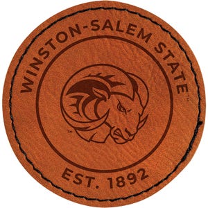 May include: A round, brown leather coaster with black stitching around the edge. The coaster features the words "WINSTON-SALEM STATE" in a circular pattern, surrounding a ram's head logo and the text "EST. 1892".