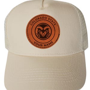 May include: Cream-colored trucker hat with a curved brim and mesh back. The front features a circular brown leather patch with the words "COLORADO STATE" and "YOUR NAME" around a ram's head logo.
