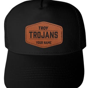 May include: A black trucker hat featuring a brown, hexagonal faux leather patch centered on the front. The patch is embossed with the text TROY, TROJANS, and YOUR NAME in black lettering, with horizontal lines separating the words.