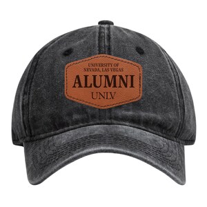 May include: A black, distressed-look baseball cap with a curved brim. A brown leather patch on the front displays the text "UNIVERSITY OF NEVADA, LAS VEGAS" above "ALUMNI" and "UNLV" in a serif font.