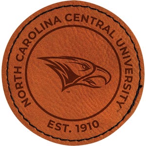 May include: A round, brown leather coaster with black stitching around the edge. The coaster features the words "NORTH CAROLINA CENTRAL UNIVERSITY" in a circular pattern, with an eagle logo in the center and "EST. 1910" below.