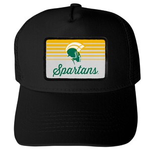 May include: A black trucker hat featuring a rectangular patch on the front. The patch displays a green and white Spartan helmet icon above the word Spartans in green script, set against a background of horizontal yellow and white stripes.