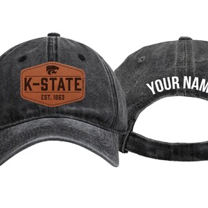 May include: A black washed-denim baseball cap featuring a brown leather-look patch on the front with the text K-STATE EST. 1863 and a wildcat logo. The back of the cap displays the white text YOUR NAME above an adjustable metal buckle strap.