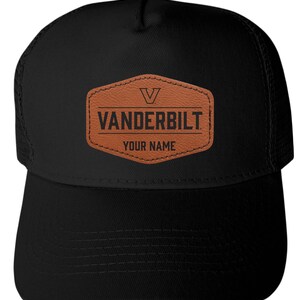 May include: A black trucker hat featuring a brown leather-like patch on the front. The patch displays a V logo, the text VANDERBILT, and the text YOUR NAME in black lettering. The hat has a solid front panel and a mesh back with a curved brim.