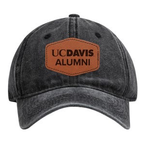 May include: A black, distressed baseball cap with a curved brim. A brown leather patch on the front displays the text "UCDAVIS ALUMNI" in a serif font. The cap has a six-panel design and an adjustable strap.