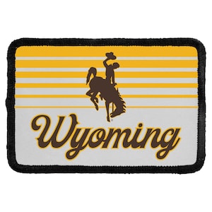 May include: A rectangular embroidered patch with a black stitched border. The design features a brown silhouette of a bucking horse and rider against a background of horizontal yellow and white stripes, with the word Wyoming in brown script.