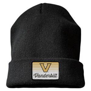May include: A black knit beanie featuring a rectangular patch on the front cuff. The patch displays a gold letter V above the word Vanderbilt in black script, set against a background of horizontal gold and white stripes on a white field.