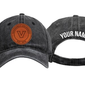 May include: A distressed black baseball cap with a leather patch featuring the Vanderbilt University logo and the text "EST. 1873." The back of the cap displays the text "YOUR NAME."