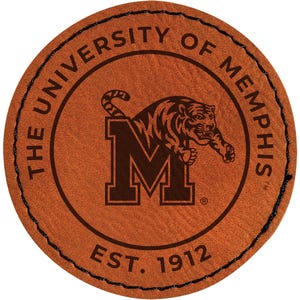 May include: A round, brown leather patch with the text "THE UNIVERSITY OF MEMPHIS" encircling a central design. The design features a tiger leaping over the letter "M" and the text "EST. 1912". The patch is stitched with black thread.