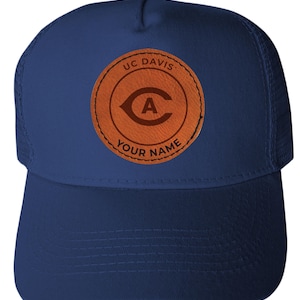 May include: A navy blue trucker hat with a curved brim and mesh back. The front features a circular brown patch with the text "UC DAVIS" and "YOUR NAME" around a stylized "C" and "A" logo.