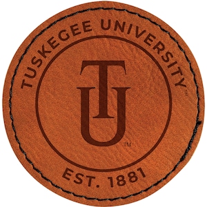 May include: A circular, brown leather patch with the words "Tuskegee University" in a circular pattern around the edge. The center features a stylized "TU" logo and the text "EST. 1881". The edge is stitched with black thread.
