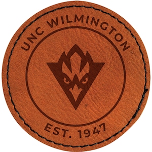 May include: A round, brown leather patch with the text "UNC WILMINGTON" in a circular pattern. Inside the circle is a brown triangle with a stylized bird design and the text "EST. 1947". The patch has a black stitched border.