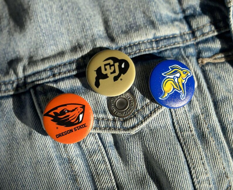 Three colorful pins or badges are attached to a denim fabric, featuring the logos of Oregon State University, the University of California, and the University of Texas.