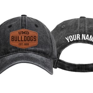 May include: A charcoal black baseball cap with a distressed finish. The front features a brown leather-like patch with the text UMD BULLDOGS EST. 1899. The back of the cap displays the white text YOUR NAME above an adjustable metal buckle strap.