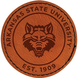 May include: A round, brown leather patch with the words "ARKANSAS STATE UNIVERSITY" in a circular pattern. The center features a wolf head logo. The text "EST. 1909" is at the bottom. The edge is stitched with black thread.