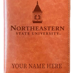 May include: A tan, textured faux leather notebook featuring the Northeastern State University logo, a clock tower icon, and the text "YOUR NAME HERE" engraved on the front cover. The notebook includes a matching brown ribbon bookmark.