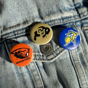 Three colorful pins or badges are attached to a denim fabric, featuring the logos of Oregon State University, the University of California, and the University of Texas.