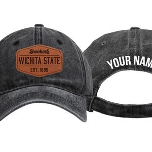 May include: A black washed-denim baseball cap featuring a brown leather-look patch on the front with the text Shockers WICHITA STATE EST. 1895. The back of the cap displays the white text YOUR NAME above an adjustable metal buckle strap closure.