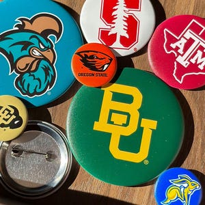 a collection of colorful pin badges, each featuring different logos and designs, including a blue pin with a bird logo, a green pin with a yellow "B" logo, and a red pin with the letters "ATM" and a map of the United States.