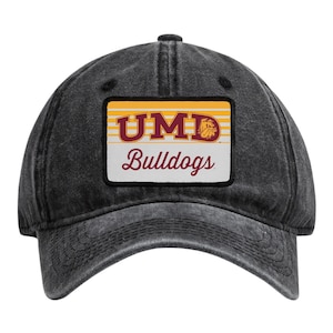 May include: A charcoal grey baseball cap with a distressed finish featuring a rectangular patch on the front. The patch displays the text UMD in maroon with a bulldog mascot icon, followed by the word Bulldogs in maroon script on a white background.