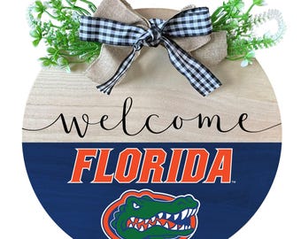 May include: A round wooden door hanger with a welcome sign and the University of Florida Gators logo. The logo is in orange and blue with an alligator in green. The top of the door hanger is decorated with a burlap bow and green floral accents.