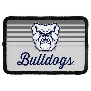 May include: A rectangular patch with a black stitched border featuring a gray and white horizontal striped background. Centered is a white bulldog head illustration with dark blue outlines. Below the graphic is the word Bulldogs in dark blue.