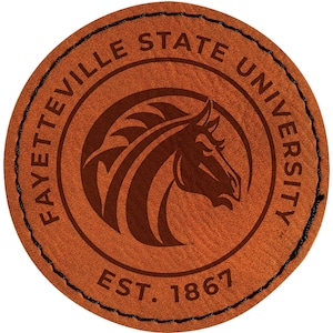 May include: A round, brown leather patch with the words "FAYETTEVILLE STATE UNIVERSITY" in a circular pattern. Inside the circle is a horse head logo. The text "EST. 1867" is at the bottom. The edge is stitched with black thread.