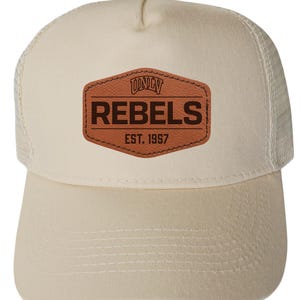 May include: A beige trucker hat with a curved brim and mesh back. The front features a brown leather patch with the text "UNLV REBELS EST. 1957" in dark brown lettering. The hat has a snap closure.
