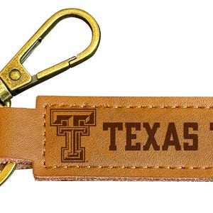 May include: A brown leather keychain with a gold metal key ring and a clip. The keychain has the Texas Tech logo and the words "TEXAS TECH" engraved on it.