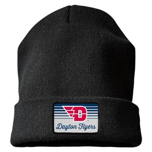 May include: A black knit beanie featuring a rectangular patch on the front cuff. The patch displays a red letter D with wings, blue horizontal stripes, and the text Dayton Flyers in a blue script font against a light gray background.