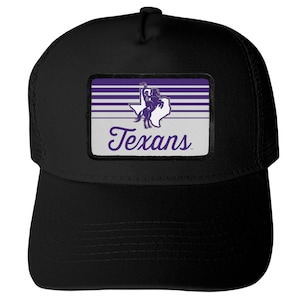 May include: A black trucker hat featuring a rectangular patch on the front. The patch displays a purple and white graphic of a horse and rider silhouette over a map of Texas, with horizontal stripes and the word Texans written in purple script.