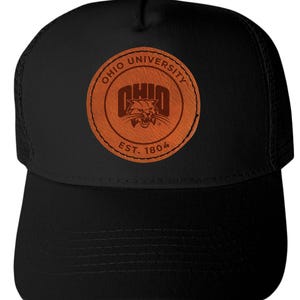 May include: Black trucker hat featuring a brown leather patch with the words "Ohio University" and "Est. 1804" encircling a graphic of a snarling bobcat. The hat has a curved brim and a mesh back.