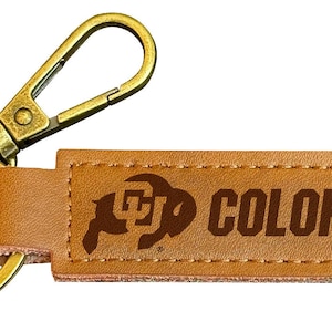 May include: A brown leather keychain with a gold metal key ring and a clip. The keychain has a brown leather tag with the University of Colorado logo and the text "COLORADO" in brown.