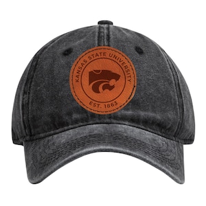 May include: A black, distressed baseball cap with a curved brim. A circular, brown leather patch features the words "Kansas State University" and "Est. 1863" around a Wildcat logo.