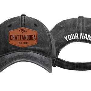 May include: A black washed-denim baseball cap featuring a brown leather-look patch on the front with the text CHATTANOOGA EST. 1886. The back of the cap displays the white text YOUR NAME and includes an adjustable metal buckle strap closure.