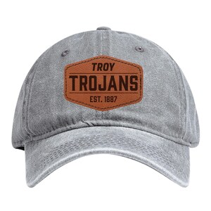 May include: A gray baseball cap with a curved brim. The cap features a brown leather patch with the words "TROY TROJANS EST. 1887" in a bold, dark font. The cap is made of a soft, worn material.