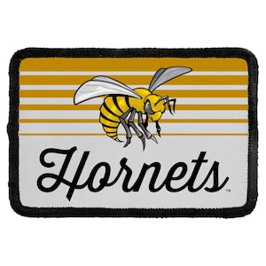 May include: A rectangular patch with a black stitched border featuring a yellow and black hornet illustration centered above the word Hornets in black script. The background consists of horizontal yellow and white stripes on the top half.
