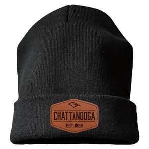 May include: A black knit beanie featuring a brown faux leather patch on the front. The patch is hexagonal and displays a bird icon above the text CHATTANOOGA and EST. 1886 in dark brown lettering. The beanie has a folded cuff design.