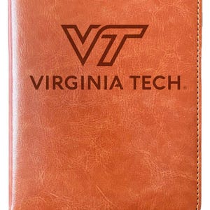May include: A brown leather journal with a stitched border and a ribbon bookmark. The front cover features a black and white logo for Virginia Tech.