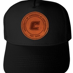 May include: Black trucker hat featuring a brown leather patch with the text "UNIVERSITY OF TENNESSEE AT CHATTANOOGA" around a large letter "C". The patch also includes "EST. 1886". The hat has a curved brim and mesh back.