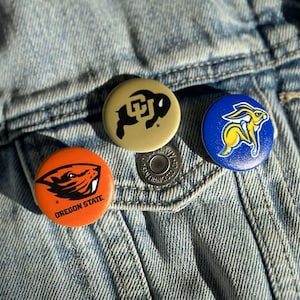 Three colorful pins or badges are attached to a denim fabric, featuring the logos of Oregon State University, the University of California, and the University of Texas.