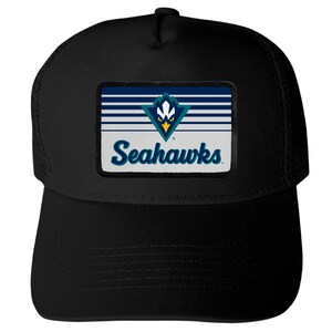 May include: A black trucker hat featuring a rectangular patch on the front. The patch displays a blue and white striped background with a stylized bird logo and the text Seahawks in blue script lettering centered below the graphic icon.