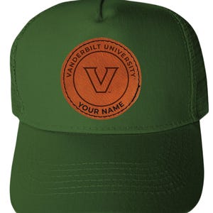 May include: Green trucker hat with a brown leather patch featuring the text "Vanderbilt University" and a "V" logo. The bottom of the patch reads "Your Name". The hat has a mesh back and a curved brim.