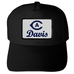 May include: A black trucker hat featuring a rectangular white patch on the front. The patch displays a dark blue logo with a stylized letter C enclosing the letter A, positioned above the dark blue text Davis in a cursive script font.