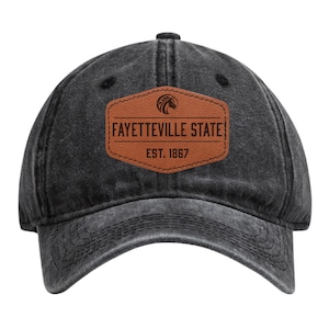 May include: A black, distressed-look baseball cap with a curved brim. The cap features a brown leather patch with the words "FAYETTEVILLE STATE" and "EST. 1867" in white lettering. A graphic of a horse is above the text.