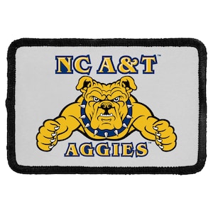 May include: A rectangular patch with a black stitched border on a light gray background. It features the text NC A&T in blue and yellow, and the text AGGIES in blue and yellow. A yellow bulldog with a blue spiked collar is centered in the design.