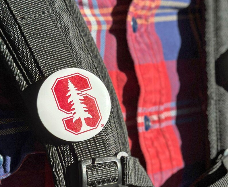 A close-up of a backpack strap with a white circular button featuring a red tree logo attached to it.